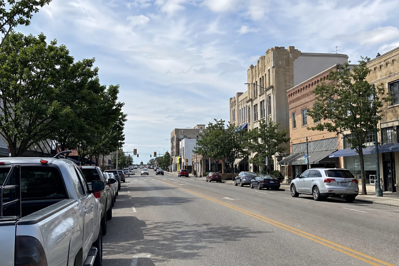 Downtown main street with brick and tan stone storefront buildings, street trees, parked pickup trucks and cars along both curbs under a high-cloud sky