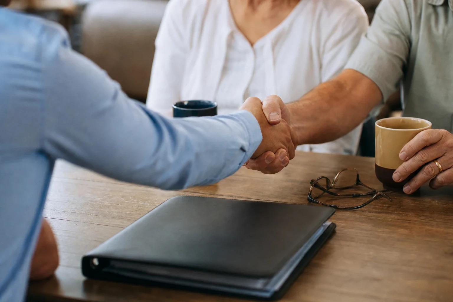 Over-the-shoulder view of an agent shaking hands with an older man at a wooden kitchen table, with a closed black folder, reading glasses and coffee mugs