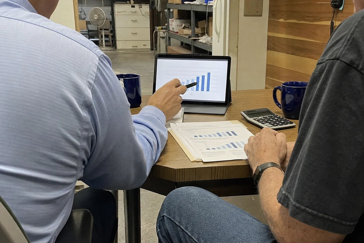 Agent in a blue shirt points a pen at a bar chart on a tablet while a business owner reviews printed charts at a small table in a shop back office